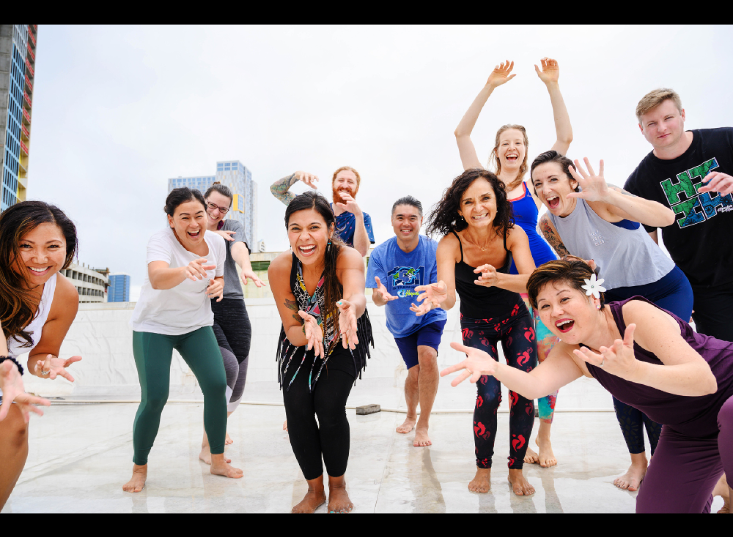 A diverse group of people joyfully participating in a movement or dance class on a rooftop, smiling and reaching forward with enthusiasm.