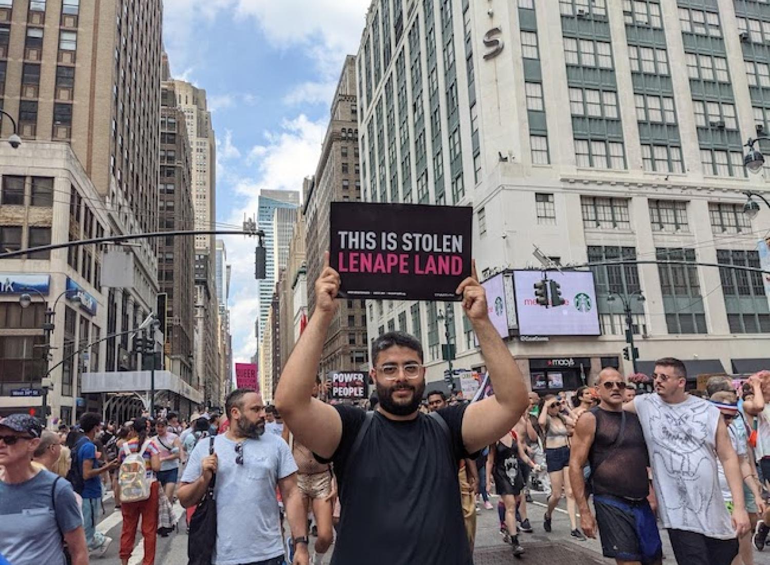 A man in a march down a city street carries a small black sign that reads "This is stolen Lenape Land" in pink and white text.
