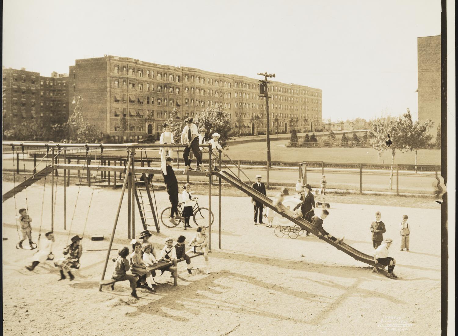 A black-and-white photograph from 1921 showing multiple children playing on a playground with swings, a slide, and a seesaw. Behind the playground is a street and apartment buildings.