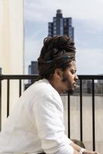 Kamau wear, un homme noir avec ses locs torsadées en chignon sur la tête, est assis de profil au premier plan. Son regard est baissé.