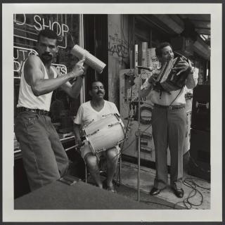 Black and white photograph of three men playing musical instruments outsde of a shop. One man is seated with a drum on his lap while the other two are standing, with one strumming a guira and another smiling as he plays his instrument.