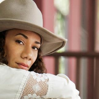 A close up portrait of Martha Redbone. She is wearing a white dress with long sleeves and a greyish tan rimmed hat. 