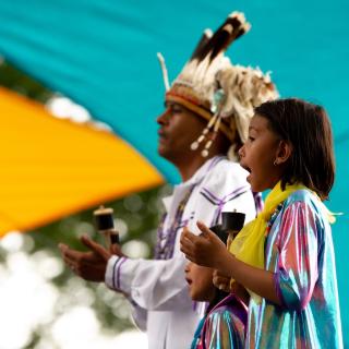 Un homme et une jeune fille en costume traditionnel chantent devant un fond turquoise.