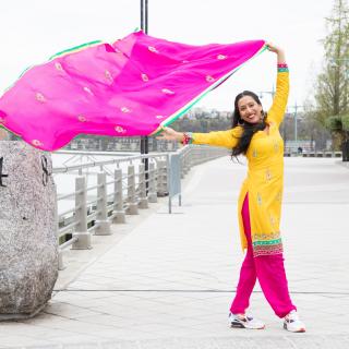 An image of a woman dressed in a bright yellow top and pink bottom holding a pink scarf that blows in the wind while smiling at the camera and standing on a boardwalk