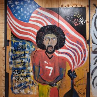 19年COVID-2020大流行和种族正义起义期间制造的胶合板艺术品。科林·卡佩尼克（Colin Kaepernick）站在前景中，弯曲的膝盖上挥舞着美国国旗。