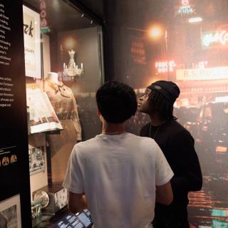 Two students stand in front of an exhibition case featuring an open book, small chandelier, and a shirt/skirt outfit. An image of an old Times Square street covers the entire wall behind them.