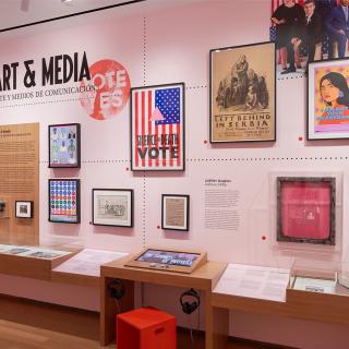 A gallery with pink walls and wooden cases showing photographs, sculptures, paintings, and interactive displays