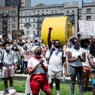 A crowd of protestors who are racially mixed but predominantly Black stand in solidarity in the plaza outside of the Brooklyn Museum. Some are holding signs, others are raising fists. All are wearing white. 