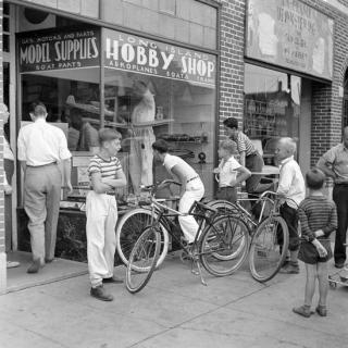 Group of youths on bicycles looking into the window of a Hobby Shop selling airplanes and train models. 