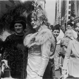 Two drag queens posing for a photo with an older woman watching