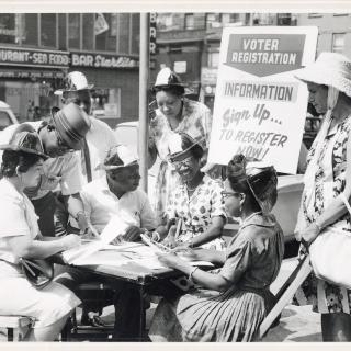 A black and white photograph of a group of Black men and women gathered around a table on the street with a sign that says Voter Registration behind them. 