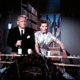 Spencer Tracy and Katharine Hepburn are standing next to each other and leaned on a rail. They are looking toward the lower right-hand corner of the screen. 