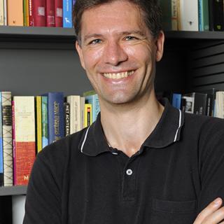 Foto de cabeza de un hombre sonriente con una camisa negra con cuello de pie con los brazos cruzados frente a una librería.