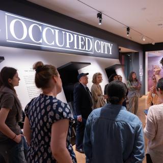 A group of visitors stands in a museum gallery beneath a large illuminated sign reading “THE OCCUPIED CITY,” listening to a curator and viewing wall-mounted exhibition panels in a modern, well-lit space.