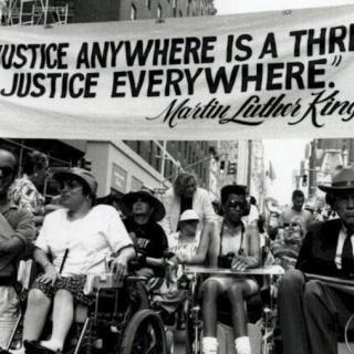 Uma multidão de pessoas com deficiência e pessoas em cadeiras de rodas se reúne sob uma faixa que diz "A injustiça em qualquer lugar é uma ameaça à justiça em todos os lugares" Martin Luther King Jr.