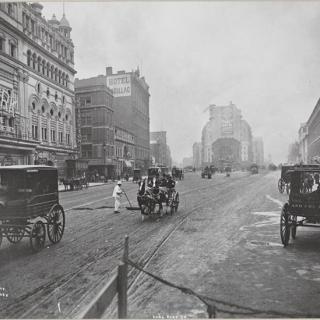 A black-and-white photograph showing the intersection of Broadway and 42nd Street, the present-day location of Times Square. The city street is surrounded by multistory buildings, and horse-drawn carriages drive in the street. In the center of the street is a New York City Sanitation worker dressed in white.