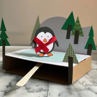 A photo of a craft project using paper and cardboard to create a penguin wearing a scarf skating on a frozen field of ice. 