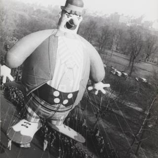 Large balloon of a clown floats about city streets, with a crowd of onlookers below. 