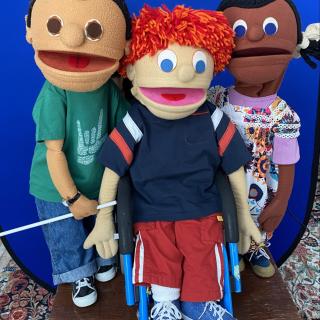 3 puppets created by NYC Kids Project standing on brown wood table, with bright blue background. One male puppet, Latinx, with dark hair, green tshirt , blue jeans , is holding a white cane. One male puppet with light skin and red hair uses a wheelchair and wears red pants, blue shirt with stripes on the arms. One female puppet, African-American has a colorful flowered dress over a lavender shirt and hair in two braids with white bows.  