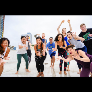 A diverse group of people joyfully participating in a movement or dance class on a rooftop, smiling and reaching forward with enthusiasm.