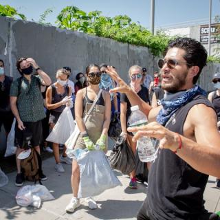 A man holding a water bottle in one hand addresses a crowd of people with bags and masks standing in front of a wooden fence.