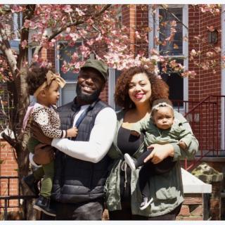 A man and woman, each holding a young child, stand smiling next to each other in front of a blossoming tree and brick building.