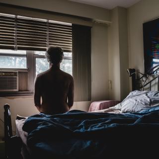 A man sits on a bed shirtless, facing his window with an AC unit installed.