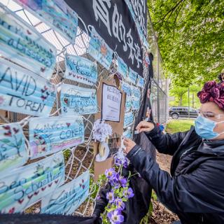 A woman places a mask with a name written on it on a fence covered in other blue face masks with names.