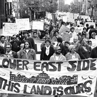 Uma foto em preto e branco de um grupo de adultos marchando por uma rua do Lower East Side. As pessoas na frente seguram uma faixa que diz: "Lower East Side. Esta terra é nossa."
