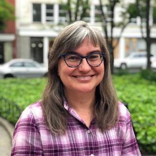 Headshot of a smiling woman wearing dark blue reading glasses and a plaid, purple shirt outdoors.