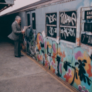 Dois homens mais velhos de terno leem jornais em uma plataforma do metrô em frente a um vagão coberto de graffiti.