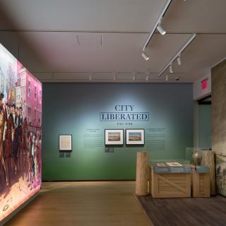 Installation view of the galleries, showing a large illuminated painting, objects in cases, and wall text saying, “City Liberated.”