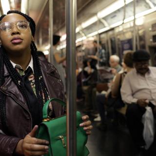 Imagem em close de mulheres de jaqueta roxa segurando uma sacola verde, sentadas em um trem do metrô de Nova York com outros passageiros.