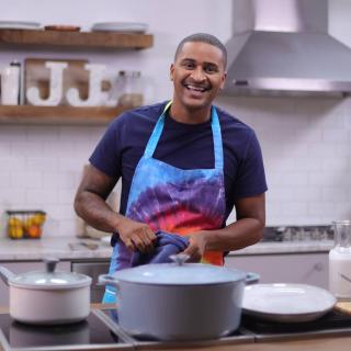 Chef JJ sorrindo em uma cozinha usando um avental tie dye arco-íris.