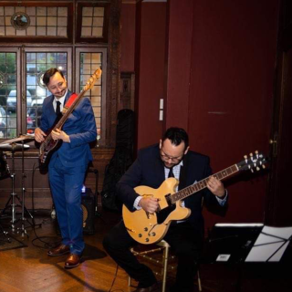 Imagen de un bajista tocando una guitarra y un guitarrista eléctrico sentado y tocando una guitarra eléctrica en una habitación con una ventana y autos al fondo de la ventana