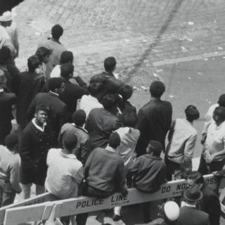 Uma visão panorâmica em preto e branco de um grupo de pessoas na rua. Barricadas policiais delineiam o espaço.
