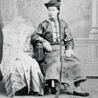 A man in a hat with a long braid sits in a chair. 