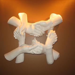 An image of four sculpted hands in white plaster interlocked and holding one another, with a soft yellow light glowing behind them.