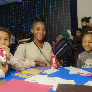 A woman and two young children smile as they make art at a table.
