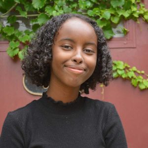 Isra Hirsi is in front of a burgundy door covered in ivy. She wears a black mock turtle neck and smiles directly at the camera. 